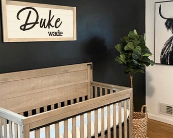 May include: A nursery room with a light wood crib, a framed black and white Highland cow print, and a sign that reads "Duke wade". The room has a black accent wall, a white wall, and a woven basket with a plant.
