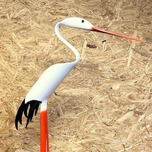 May include: White bird sculpture with a long orange beak and black and white tail feathers. The sculpture is standing on a brown surface.