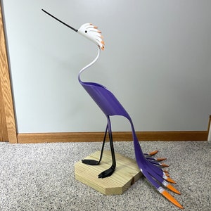 May include: A purple and white metal sculpture of a bird with an orange and white crest. The bird has long, thin legs and a long, thin tail. The sculpture is standing on a wooden base.