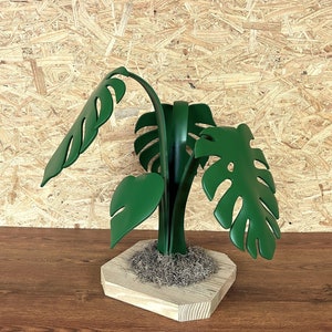 May include: A green metal sculpture of a plant with large leaves. The sculpture is on a wooden base with a brown surface.