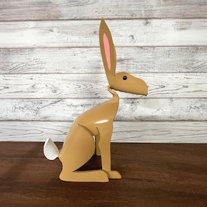 May include: A light brown wooden hare sculpture in a seated pose. The hare features pink inner ears and a white tail. The minimalist design is set against a wood-panelled background. The sculpture is a decorative item.