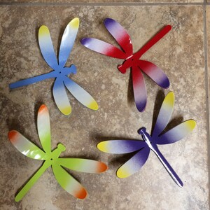 May include: Four metal dragonfly wall decor pieces in different colors. The dragonflies are painted in shades of blue, red, green, and purple with white accents.