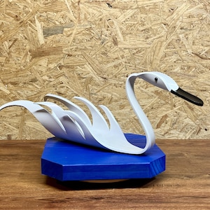 以下が含まれることがあります： 黒い嘴と湾曲した首を持つ白い白鳥の彫刻。 白鳥は青い六角形の木製の台座に取り付けられています。 彫刻は木目調の背景に配置されています。