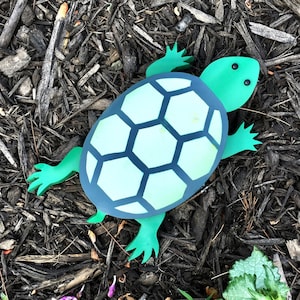 May include: A green and blue turtle-shaped garden decoration with a textured shell. The turtle is lying on a bed of brown mulch.