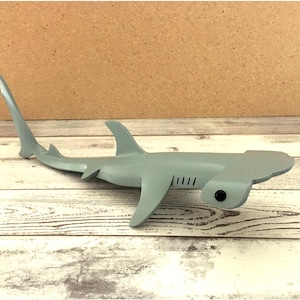 May include: A gray hammerhead shark figurine with a flat head and a long, slender body. The shark has a textured surface and a realistic appearance.