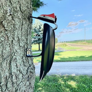 May include: A black metal woodpecker sculpture with red and white accents is attached to a tree trunk. The sculpture is facing to the right and is partially visible. The background is a green grassy field with a road and a blue sky with white clouds.