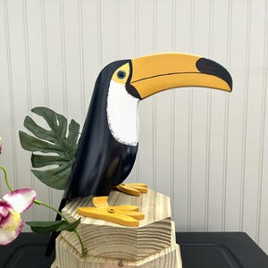 May include: A wooden toucan sculpture with a yellow beak, black body, and white accents. The toucan is perched on a wooden base with a leafy green plant behind it.
