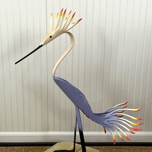 May include: A wooden bird sculpture with a long neck, a long beak, and a long tail. The bird is painted in shades of purple, white, and orange. The bird is standing on a wooden base.