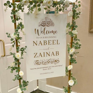 May include: A white rectangular wedding welcome sign with gold lettering featuring the names Nabeel and Zainab, Arabic calligraphy, and the text And He placed between you love and mercy, displayed on a gold metal frame adorned with white roses and eucalyptus.