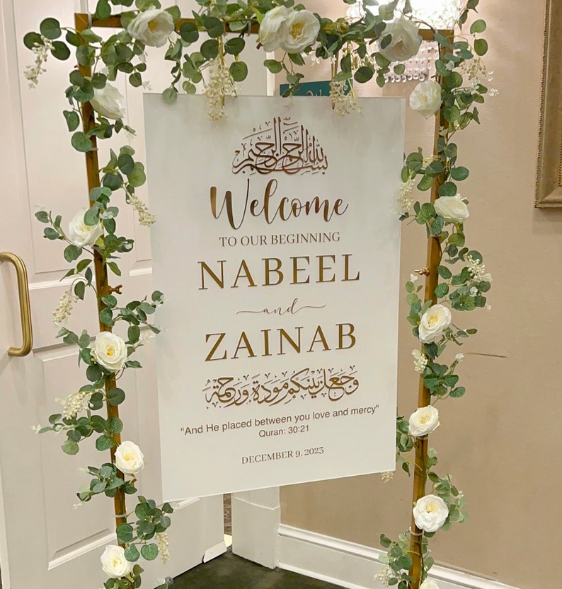 May include: A white rectangular wedding welcome sign with gold lettering featuring the names Nabeel and Zainab, Arabic calligraphy, and the text And He placed between you love and mercy, displayed on a gold metal frame adorned with white roses and eucalyptus.
