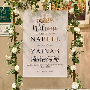 May include: A white rectangular welcome sign with gold and black text featuring the names Nabeel and Zainab, Quranic calligraphy, and the date December 9, 2023. The sign is displayed on a gold metal frame decorated with white roses and green leaves.