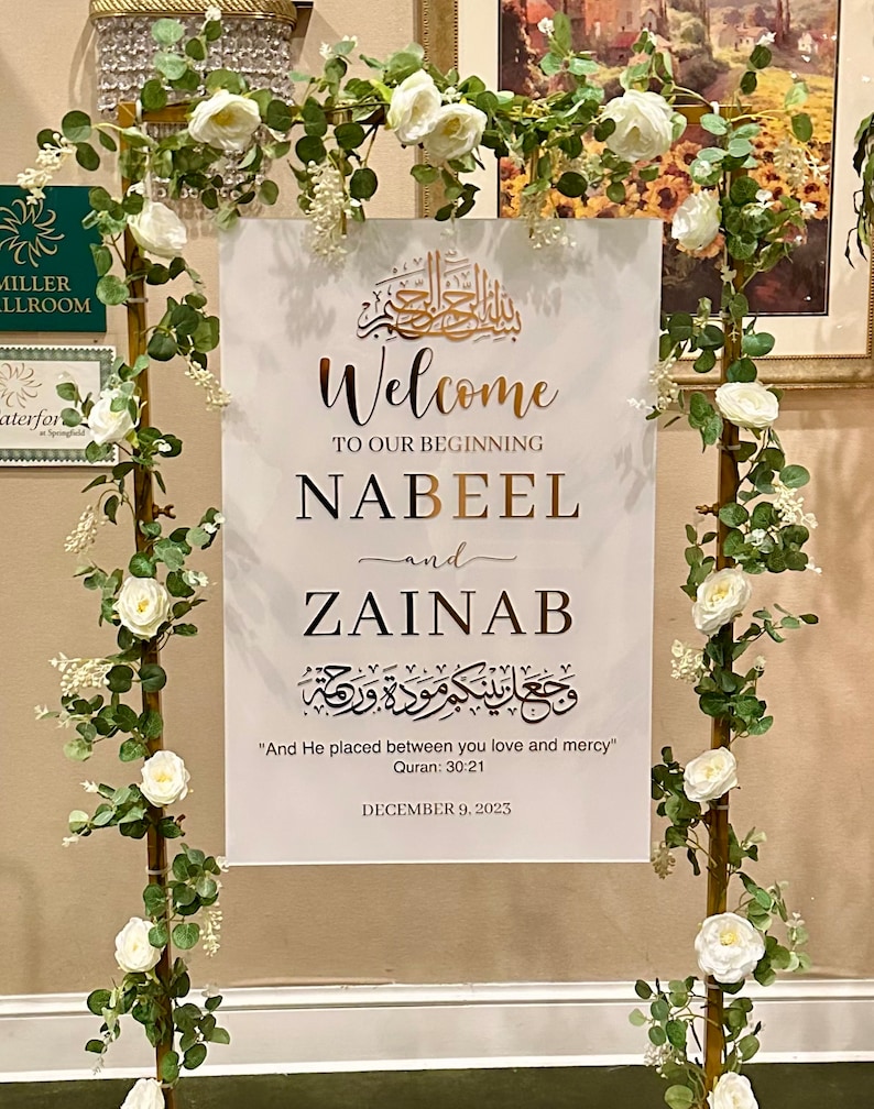 May include: A white rectangular welcome sign with gold and black text featuring the names Nabeel and Zainab, Quranic calligraphy, and the date December 9, 2023. The sign is displayed on a gold metal frame decorated with white roses and green leaves.
