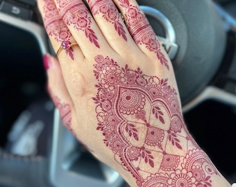 May include: A close-up of a hand with intricate henna designs in a deep red color. The henna is applied to the back of the hand, fingers, and wrist. The henna design features a variety of patterns, including circles, swirls, and leaves.