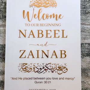 May include: A white rectangular sign featuring gold lettering that reads Welcome to our beginning, Nabeel and Zainab, along with Arabic calligraphy, the English translation And He placed between you love and mercy, Quran 30:21, and December 9, 2023.