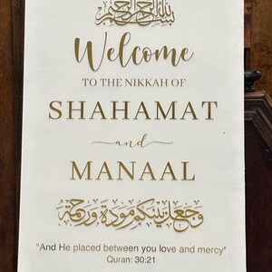 May include: A rectangular white wedding welcome sign featuring gold lettering. The text reads: Welcome to the Nikkah of Shahamat and Manaal, with Arabic calligraphy, the Quranic verse 30:21, and the date December 26, 2023, in a clean font.