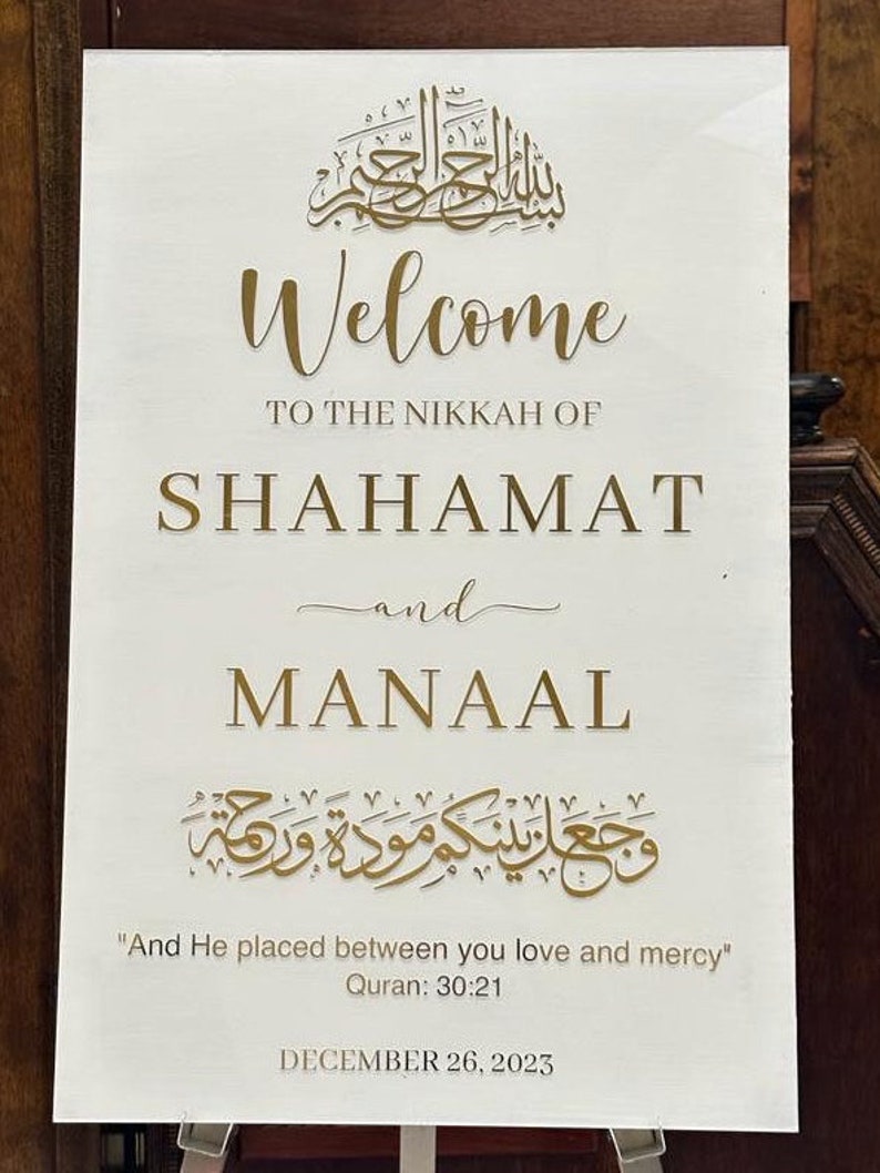 May include: A rectangular white wedding welcome sign featuring gold lettering. The text reads: Welcome to the Nikkah of Shahamat and Manaal, with Arabic calligraphy, the Quranic verse 30:21, and the date December 26, 2023, in a clean font.