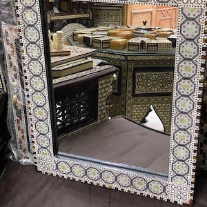 Può includere: Uno specchio decorativo con una cornice a motivi geometrici bianchi e neri. Lo specchio riflette una stanza con mobili in legno e intarsiati con madreperla.