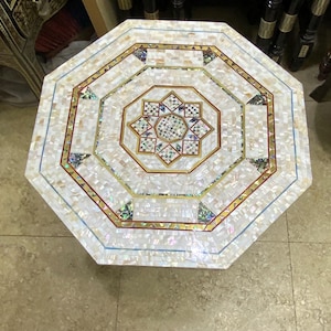 Op de afbeelding: Een achthoekige mozaïektafel met een parelmoer oppervlak. De tafel heeft een centraal sterrenpatroon en gouden en iriserende accenten. De afmetingen van de tafel zijn niet zichtbaar op de afbeelding.