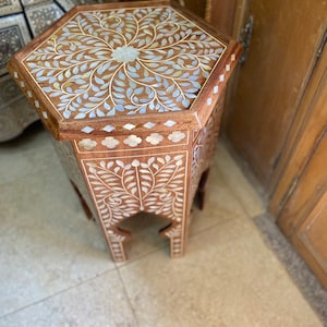 May include: A hexagonal wooden side table with intricate floral carvings inlaid with mother-of-pearl. The table has a delicate, ornate design and a light brown finish.