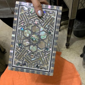 Könnte beinhalten: Eine rechteckige Schachtel mit einer dekorativen Einlage aus Perlmutt und schwarzen geometrischen Mustern. Die Schachtel ist aus Holz und hat eine glänzende Oberfläche.