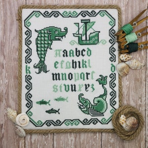May include: A white cross-stitch embroidery with a green border. The embroidery features a green alphabet, a green dragon, a green ship, and two green fish. The embroidery is framed with a brown rope.