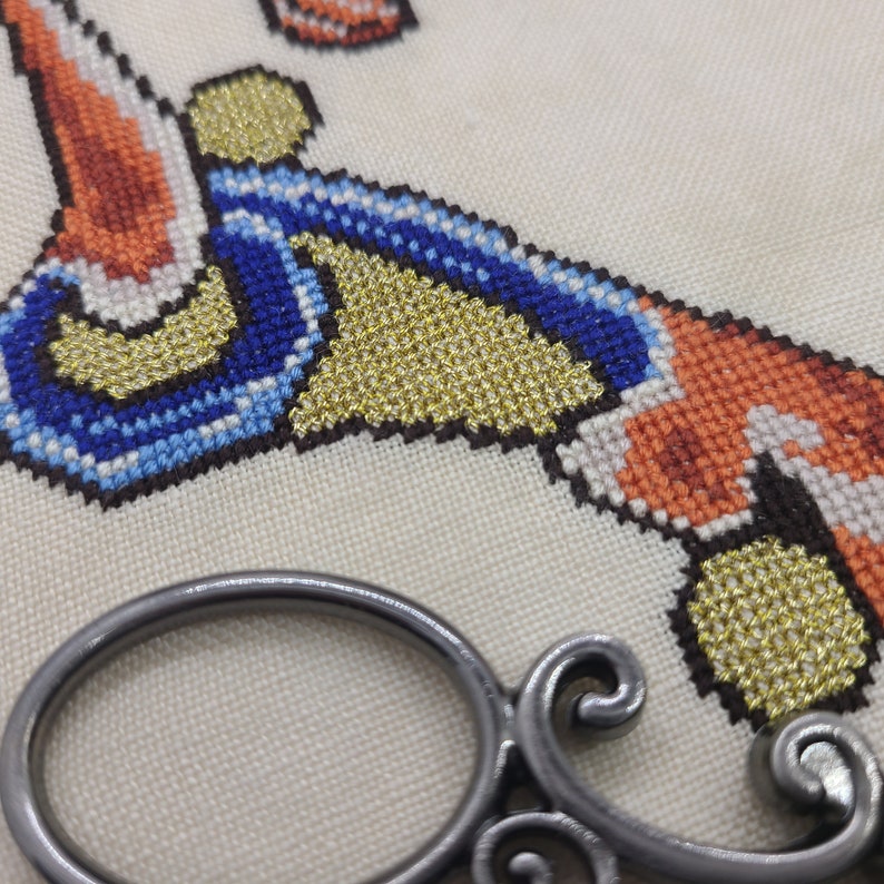May include: Close-up of a cross-stitch embroidery project featuring a geometric pattern in shades of blue, orange, gold, and white. The fabric is a natural linen color.