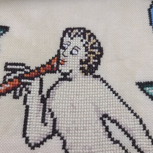 May include: A cross-stitch embroidery of a person playing a musical instrument. The person is depicted in a white tunic with brown hair and is holding a red and brown instrument to their lips.