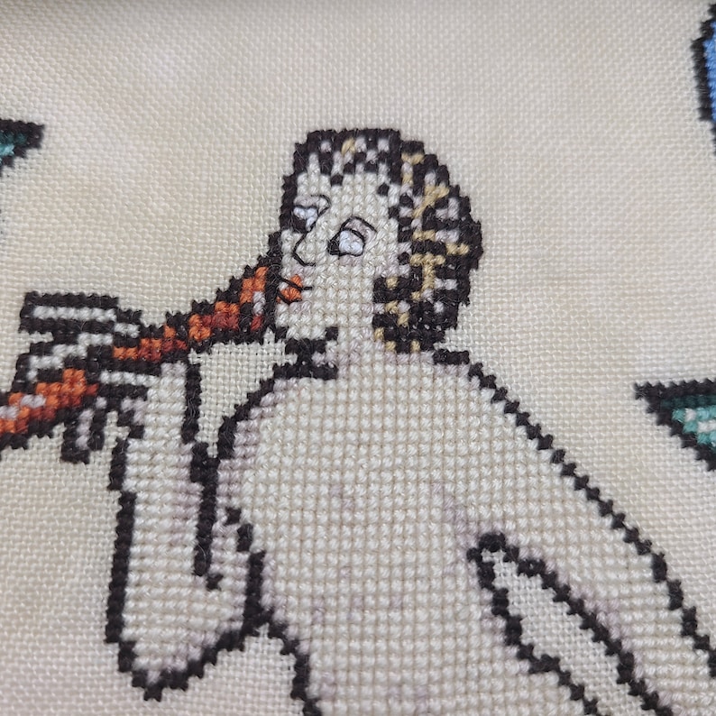 May include: A cross-stitch embroidery of a person playing a musical instrument. The person is depicted in a white tunic with brown hair and is holding a red and brown instrument to their lips.