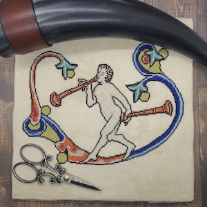 May include: A cross-stitch embroidery of a medieval-style figure playing a musical instrument. The figure is depicted in a heart-shaped frame with intricate floral designs. The embroidery is done on a beige linen fabric.