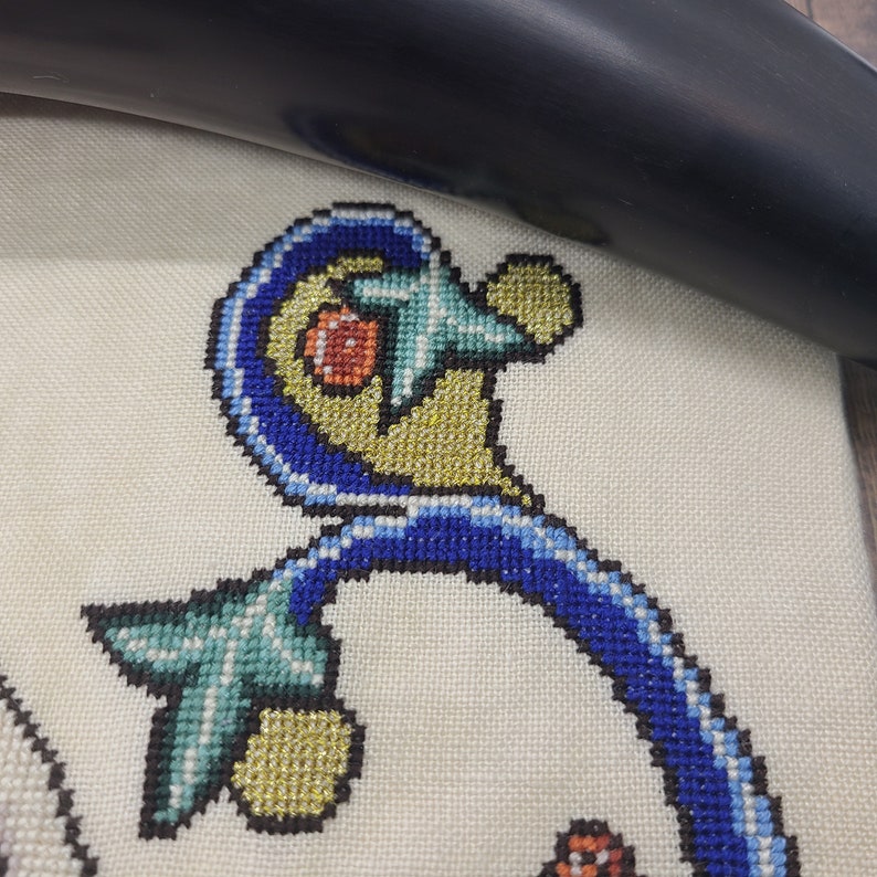 May include: Close-up of a blue, white, and gold cross-stitch design featuring a stylized floral pattern.