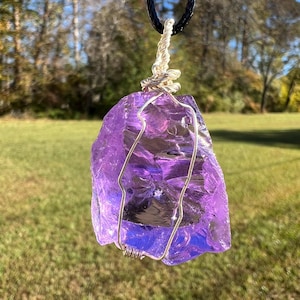 May include: A raw, purple amethyst pendant necklace. The amethyst is wrapped in silver-colored wire and suspended from a black cord. The gemstone has a rough, natural texture and is displayed against a blurred outdoor background.