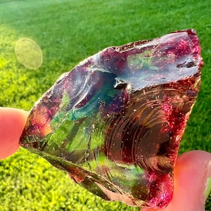 May include: A piece of iridescent glass with a mix of red, green, and blue hues. The glass has a rough, textured surface and is held up against a background of green grass.