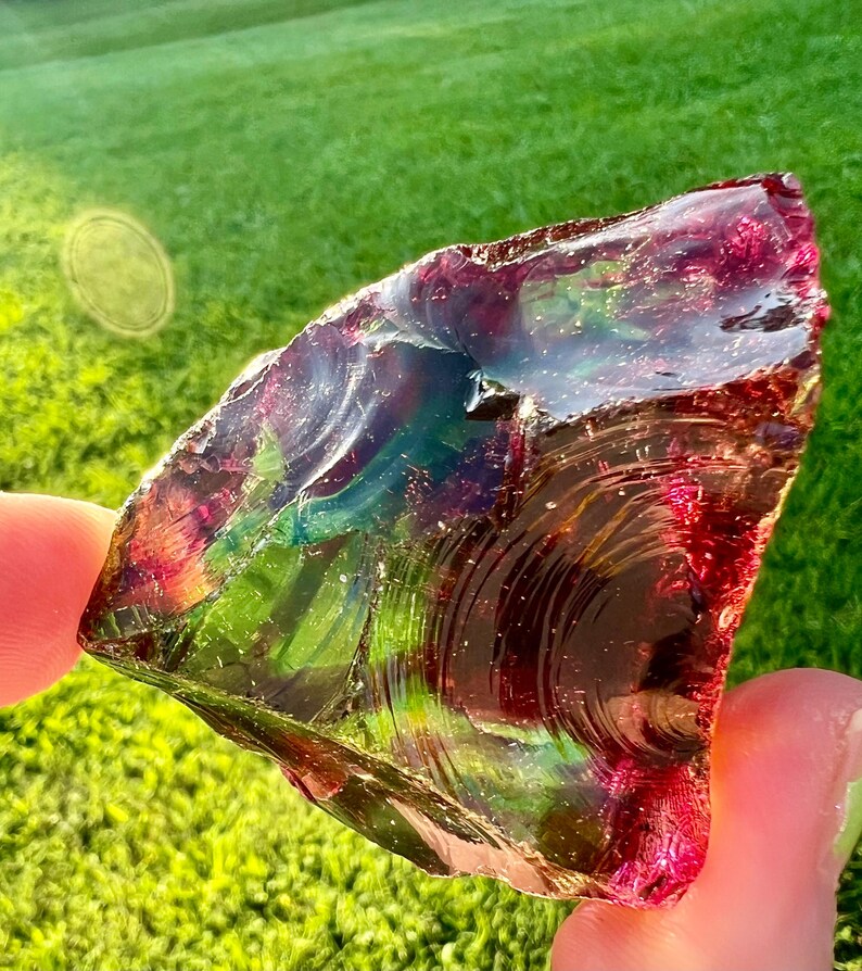 May include: A piece of iridescent glass with a mix of red, green, and blue hues. The glass has a rough, textured surface and is held up against a background of green grass.