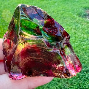 May include: A large, irregularly shaped piece of glass with a polished, iridescent surface. The glass displays a range of colors, including deep reds, greens, and hints of gold. The object is held against a backdrop of green grass.