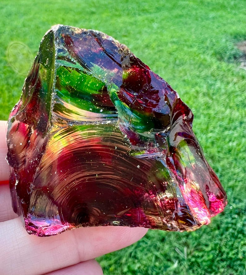 May include: A large, irregularly shaped piece of glass with a polished, iridescent surface. The glass displays a range of colors, including deep reds, greens, and hints of gold. The object is held against a backdrop of green grass.