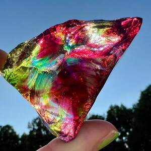 May include: A vibrant, iridescent glass shard, held between fingers, displays a spectrum of colors including red, green, and yellow. The sunlight catches the textured surface, creating a dazzling display against a blue sky.