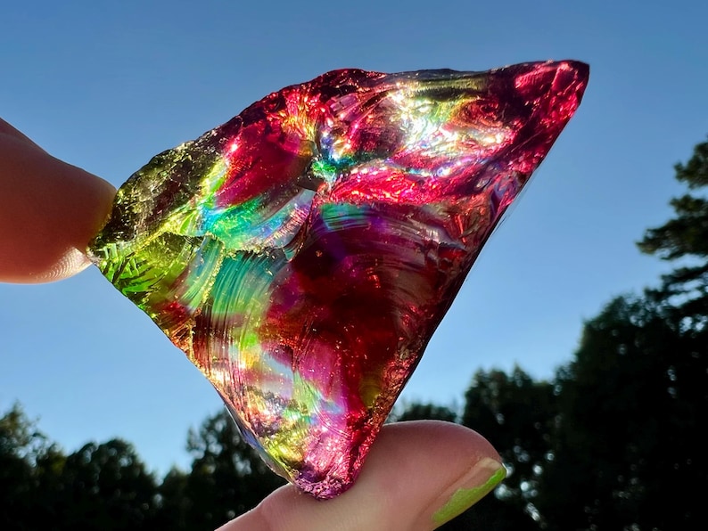 May include: A vibrant, iridescent glass shard, held between fingers, displays a spectrum of colors including red, green, and yellow. The sunlight catches the textured surface, creating a dazzling display against a blue sky.