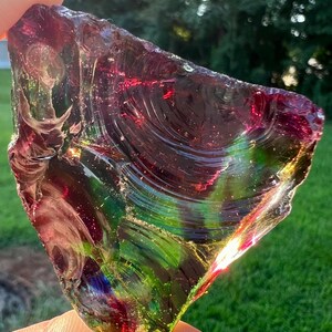 May include: A triangular piece of iridescent glass with a mix of colors, including red, green, and purple. The glass has a textured surface and is held up by a hand. The background is a blurred view of green grass and trees.