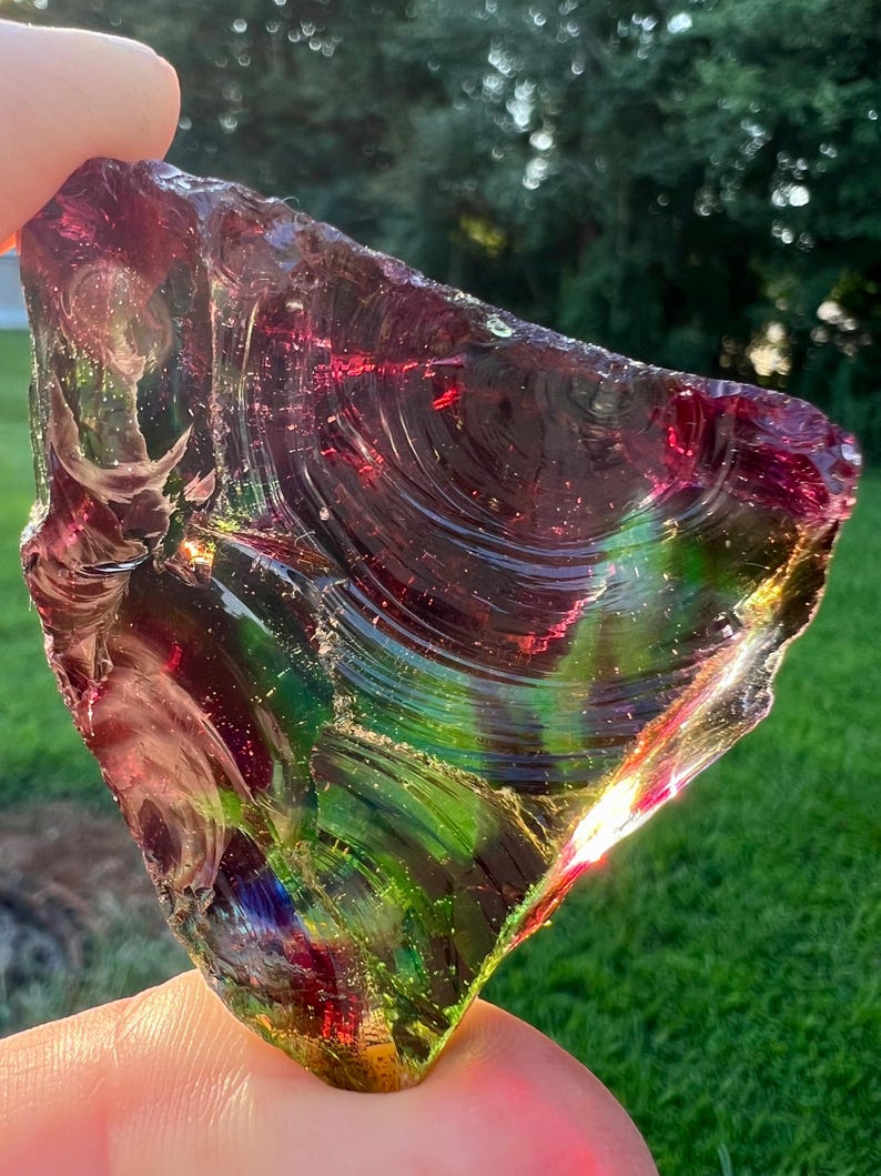 May include: A triangular piece of iridescent glass with a mix of colors, including red, green, and purple. The glass has a textured surface and is held up by a hand. The background is a blurred view of green grass and trees.