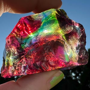 May include: A vibrant, iridescent glass rock fragment, displaying a spectrum of colors including red, green, and purple. The sunlight catches the textured surface, creating a rainbow effect. The object is held up against a blue sky.