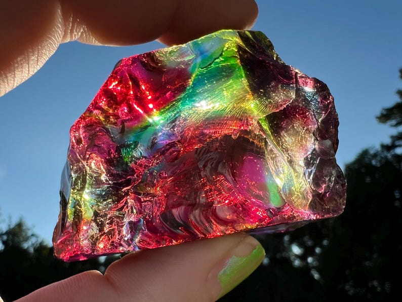May include: A vibrant, iridescent glass rock fragment, displaying a spectrum of colors including red, green, and purple. The sunlight catches the textured surface, creating a rainbow effect. The object is held up against a blue sky.
