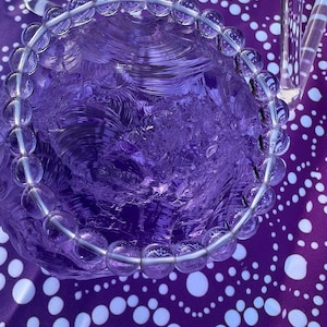 May include: A clear quartz crystal bead bracelet. The bracelet is composed of round, transparent beads strung together on a silver-colored wire. The bracelet is resting on a textured, purple glass object, with a purple and white polka dot background.
