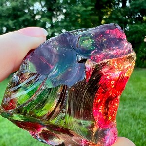 May include: A colorful, iridescent glass rock with a mix of red, green, blue, and purple hues. The rock has a rough, textured surface and is held up against a blurred green background.