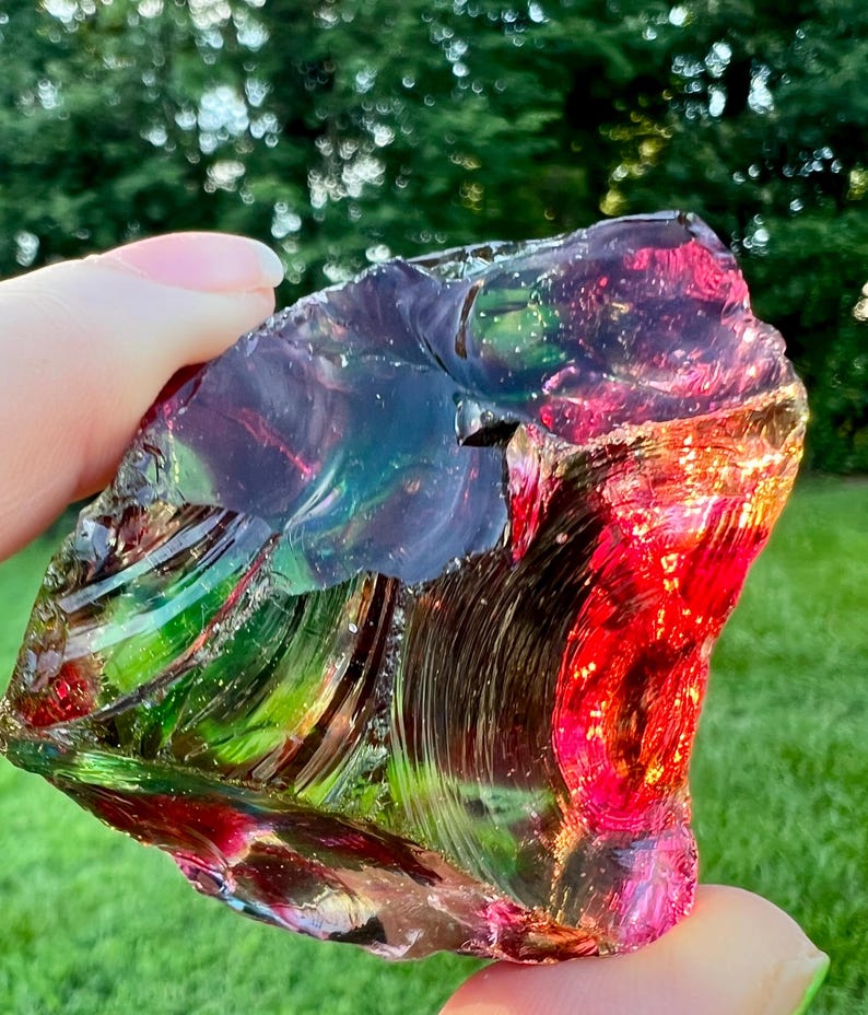 May include: A colorful, iridescent glass rock with a mix of red, green, blue, and purple hues. The rock has a rough, textured surface and is held up against a blurred green background.