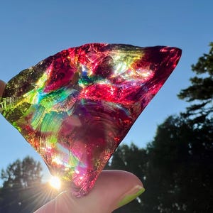 May include: A vibrant, iridescent piece of glass, held up against a bright blue sky. The glass shard displays a spectrum of colors, including red, green, and purple, with light reflecting through it. The object is irregularly shaped and appears to be a decorative item.