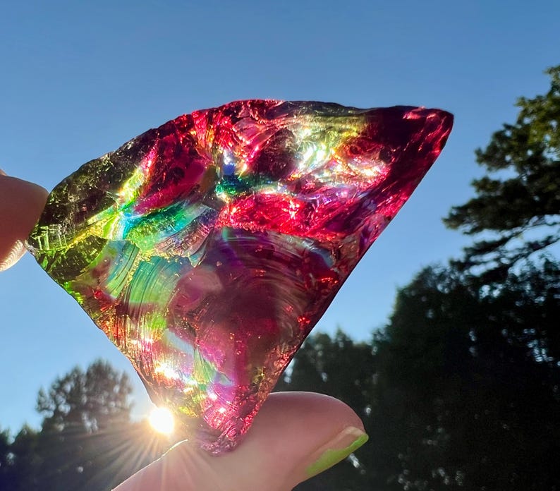 May include: A vibrant, iridescent piece of glass, held up against a bright blue sky. The glass shard displays a spectrum of colors, including red, green, and purple, with light reflecting through it. The object is irregularly shaped and appears to be a decorative item.