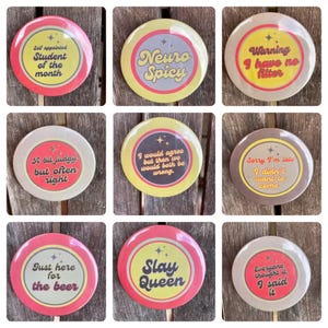 May include: Nine retro-style button badges with various sayings. The badges are round and have a red, yellow, or brown background with black and white text. The sayings include "Self appointed Student of the month", "Neuro Spicy", "Warning I have no filter", "A bit judgy but often right", "I would agree but then we would both be wrong", "Sorry I'm late I didn't want to come", "Just here for the beer", "Slay Queen", and "Everyone thought it. I said it."