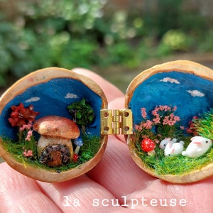 Op de afbeelding: Een miniatuur boslandschap in een walnootschelp, met een paddenstoelhuis, een blauwe lucht en twee witte konijnen. De schelp is geschilderd met een blauwe lucht en wolken, en de scène is gedetailleerd met miniatuur bomen, bloemen en gras.