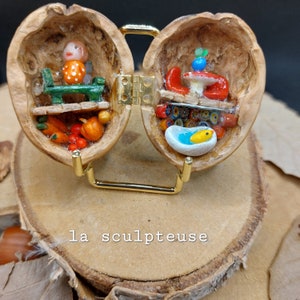 Op de afbeelding: Een miniatuur scène gemaakt in twee helften van een walnootschelpen. De linkerzijde toont een klein figuurtje met een rood-groene outfit die op een groene bank zit. De rechterzijde toont een miniatuur badkuip met een gele eend erin. De walnootschelpen zijn bevestigd aan een gouden metalen standaard.