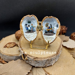 Peut inclure: Une œuvre d'art miniature en diorama présentée sur un support doré. L'œuvre est logée dans une coquille de noix, divisée en deux moitiés, chacune représentant une petite pièce avec des figurines et des objets miniatures. Le texte "La sculpteuse" est visible.
